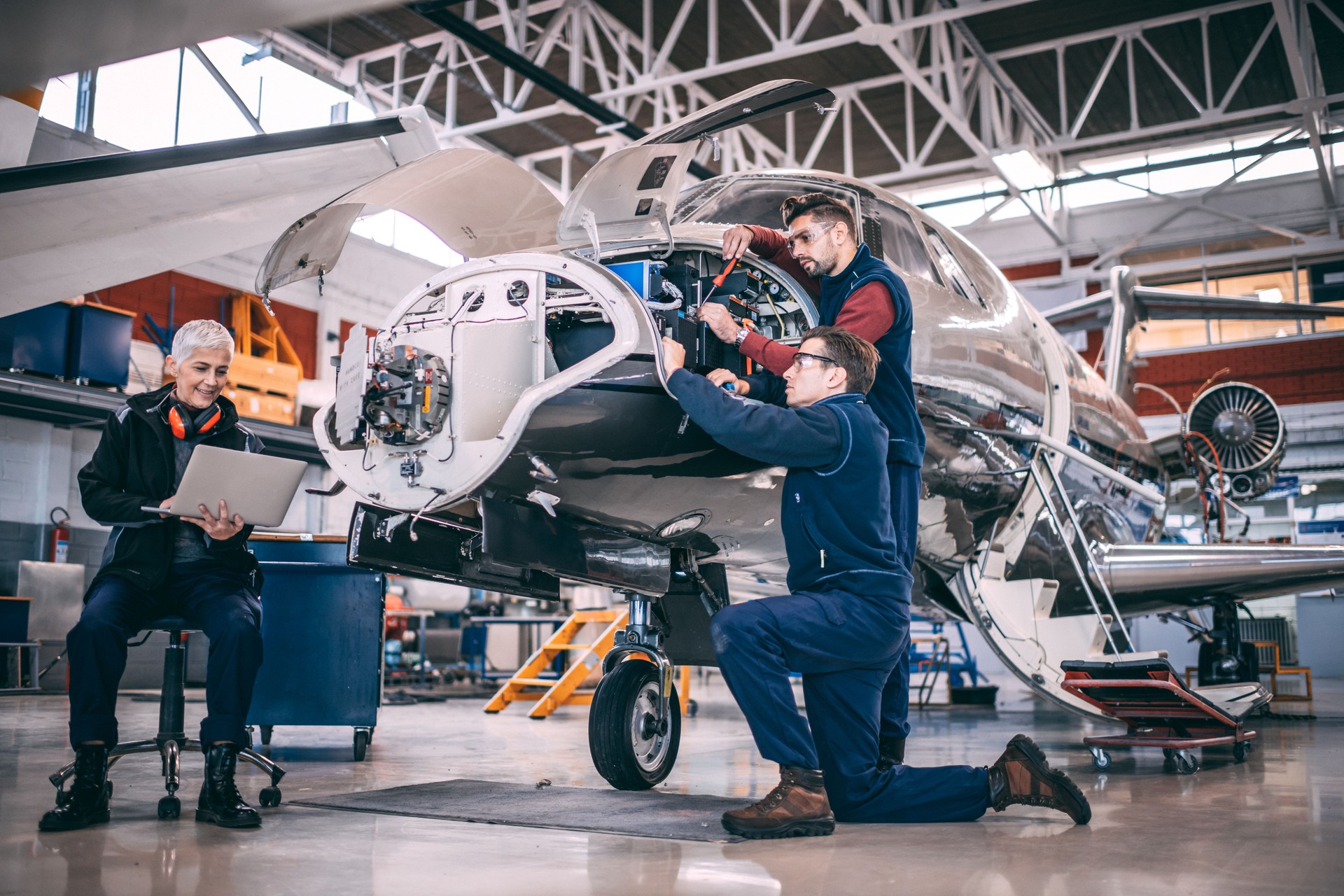 Senior female aircraft mechanic sitting and using her laptop helping her colleagues who are using hand tools to work on a small airplane in a hangar